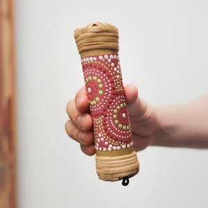May include: A hand holds a red, white, and green painted wooden tube with a woven bamboo top and bottom. The tube is decorated with a pattern of dots and circles.