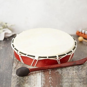 May include: A red wooden frame drum with a white head and a brown wooden beater. The drum is on a wooden surface.