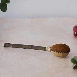 Op de afbeelding: Een bruine leren rammelaar met een houten handvat. De rammelaar is vastgebonden met een wit touwtje.