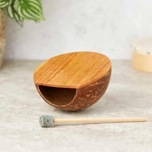Könnte beinhalten: Ein hölzernes Percussion-Instrument aus einer Kokosnussschale mit einer Holzplatte und einem kleinen Holzschlägel mit Filzspitze.
