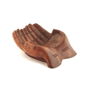 May include: A wooden hand-shaped bowl carved from dark brown wood. The bowl is shaped like two cupped hands, with detailed carvings on the fingers and palms.