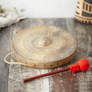 Könnte beinhalten: Ein Messinggong mit einem zentralen, erhabenen Knauf und einem Seilgriff. Der Gong wird von einem roten Schlägel mit Holzgriff begleitet. Der Gong hat ein kreisförmiges Design mit kleinen, gleichmäßig verteilten Vertiefungen.