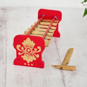 May include: A red wooden xylophone with gold painted floral designs and a wooden mallet. The xylophone has a series of wooden bars that are held in place by red strings.