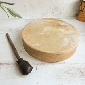 May include: A round, light brown drum with a natural skin head and a wooden frame. A dark brown drumstick with a leather-covered head rests beside the drum. The drum is set against a white wooden background.