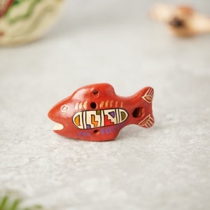May include: A red clay fish whistle with colourful geometric designs painted on it. The whistle has four holes and a small opening on the side for blowing.