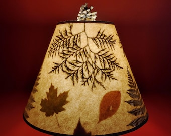 May include: A lampshade with a nature-inspired design. The beige shade features brown leaf and fern silhouettes. A small pine cone sits atop the shade. The lampshade has a black trim and is set against a red background.