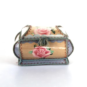 Peut inclure: Petite boîte décorative ornée de roses. Le fond est beige clair avec des roses roses et des feuilles vertes. Les bords sont bordés d'une corde bleue et verte, et une houppe pend du dessus.