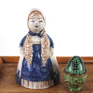 Könnte beinhalten: Eine Keramikfigur einer Frau in einem blau-weißen Kleid mit Häubchen und Zöpfen und ein grün-schwarzes Ei in einem Halter sitzen auf einem Holztablett. Die Figur hat einen ruhigen Ausdruck.