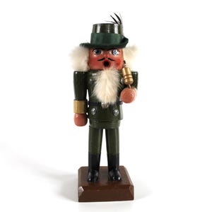 May include: A wooden nutcracker figurine in a green uniform with a matching hat and white fur trim. The figurine has a red face, black boots, and holds a gold-coloured bell. It stands on a brown rectangular base.