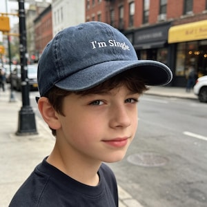 May include: A navy blue denim baseball cap with the text "I'm Single" in white. The cap features a curved peak and an adjustable strap. The cap is worn by a person.