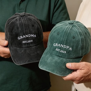 Op de afbeelding: Twee baseballpetten, één zwarte en één groene, worden getoond. De zwarte pet heeft de tekst "GRANDMA EST.2025" in het wit. De groene pet heeft de tekst "GRANDPA EST.2025" in het wit. Beide petten zijn gemaakt van een zacht, getextureerd materiaal.