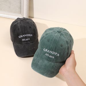 May include: Two baseball caps are shown, one black and one green. The black cap has "GRANDMA EST.2025" embroidered in white. The green cap has "GRANDPA EST.2025" embroidered in white. Both caps are made of a soft, worn fabric.