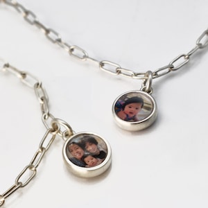 Op de afbeelding: Twee zilveren kettingen met ronde fotobedels. Een bedel heeft een foto van een baby, en de andere bedel heeft een foto van een gezin van drie.