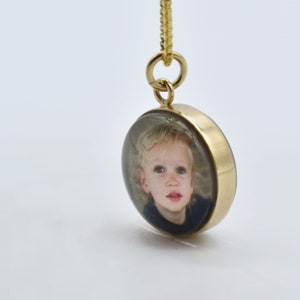 May include: Gold pendant necklace with a round glass locket featuring a photo of a young child with blonde hair and blue eyes.