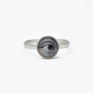 May include: A silver-toned ring featuring a circular setting with a black and white photographic print of a human eye. The band has a brushed metal finish and the central eye design is encased under a smooth, clear, rounded glass cabochon.