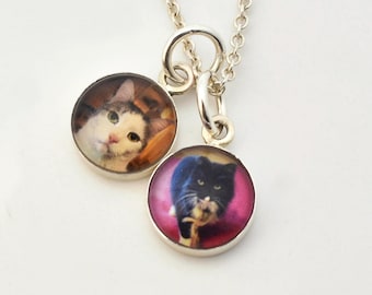 May include: A silver necklace featuring two round pendants with cat portraits. One pendant displays a white and gray cat, the other a black and white cat, both set against a pink background. The necklace has a delicate chain.