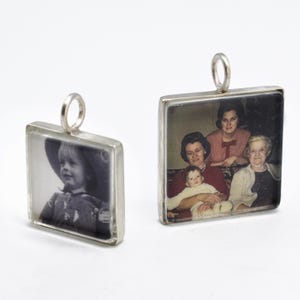May include: Two square pendants with silver frames. One pendant features a black and white photo of a child wearing a hat. The other pendant displays a color photo of a family. Both pendants have a small silver loop at the top.