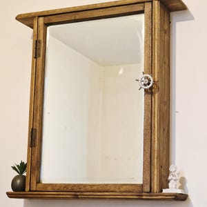 May include: A rectangular wooden wall cabinet with a mirrored door, featuring a small metal ship wheel handle. The cabinet has a top shelf and a bottom shelf, with a small dark succulent pot on the left and a white angel figurine on the right.