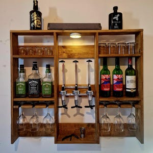 Op de afbeelding: Een houten barkast met meerdere planken, met flessen sterke drank, wijnglazen en borrelglazen. De kast heeft ingebouwde drankdispensers en een kleine lamp. Verschillende flessen sterke drank en wijn worden getoond.