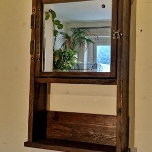 Può includere: Un mobiletto da bagno rustico in legno con anta a specchio e un piccolo ripiano. L'armadietto è tinto di marrone scuro e presenta cerniere e un fermo in ottone. Lo specchio riflette delle piante, suggerendo un'estetica naturale o vintage.