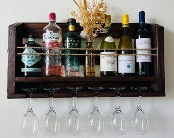 May include: A dark brown wooden wall-mounted wine rack with six glass holders and a shelf for bottles. The rack is holding six bottles of wine and spirits, including Gordon's Gin, Mermaid Gin, La Vieille Ferme, Macon-Chardonnay, and Comine Vendange.