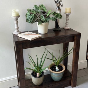 May include: A dark brown wooden console table with two shelves. The top shelf has a white ceramic candle holder, a notebook, and a pencil. The bottom shelf has two potted plants. A decorative statue of a woman is on the top shelf.