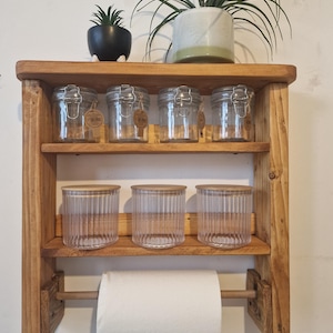 Könnte beinhalten: Rustikales Holzregal mit zwei Ebenen, das sechs Gläser mit Metallverschlüssen und drei geriffelte Gläser mit Holzdeckeln enthält. Darunter hängt eine Rolle weißer Küchenrolle. Oben stehen zwei Topfpflanzen.