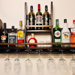 Op de afbeelding: Een donkerhouten wandrek voor wijn met planken en glazenhouders. Het rek is gevuld met verschillende flessen sterke drank, waaronder wijn, whisky en likeur. De flessen zijn in twee rijen op de planken gerangschikt, met de glazenhouders eronder hangend. Het rek is een geweldige manier om uw favoriete dranken op te bergen en te presenteren.