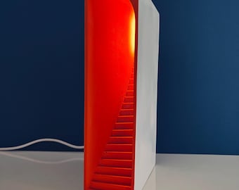 May include: A modern, rectangular lamp with a white exterior and a vibrant red interior. The interior features a staircase design that is illuminated, creating a warm glow. The lamp is placed on a white surface against a dark blue background.