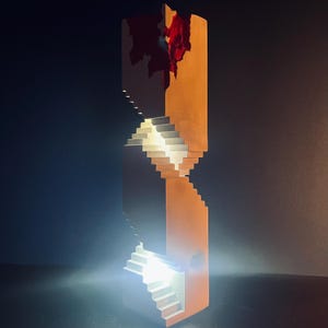Puede incluir: Una escultura abstracta con escaleras iluminadas dentro de una estructura naranja. La parte superior tiene una zona roja dentada. El diseño crea una ilusión visual de escaleras ascendentes y descendentes.