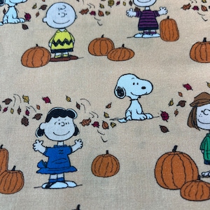 Może przedstawiać: Beżowa tkanina z postaciami z kreskówek, dyniami i opadającymi liśćmi. Snoopy, Charlie Brown, Lucy i inne postacie z Peanuts są przedstawione z pomarańczowymi dyniami. Projekt ma jesienny motyw.