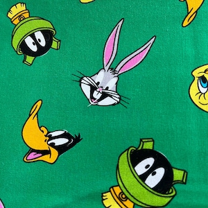 May include: A green fabric featuring cartoon characters including Bugs Bunny, Daffy Duck, Tweety Bird, and Marvin the Martian. The characters are in various poses and are set against a solid green background. The fabric is likely for crafting or apparel.