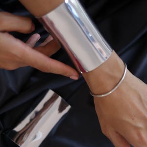 May include: A wide, polished silver-tone cuff bracelet with a smooth, reflective surface. The cuff features a minimalist, open-ended design that wraps around the wrist. A thin, simple silver-tone bangle and a small ring are also visible on the hand.