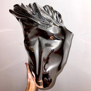 May include: A silver metal vase with a unique, abstract, and organic shape. The vase is smooth and reflective, with a flowing, fluid design.