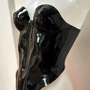 May include: A strapless, form-fitting corset top in a glossy black finish. The garment features a sweetheart neckline with moulded bust cups and a smooth, reflective surface that contours to the torso, extending down to a pointed hemline.