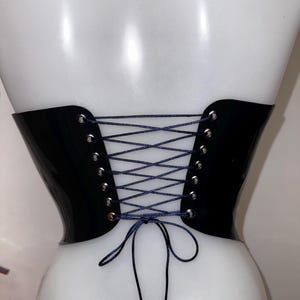 May include: A black corset belt with a glossy finish, featuring silver-toned metal eyelets along the back edges. The garment is secured with a thin, dark blue cord laced in a crisscross pattern and tied in a bow at the bottom centre.