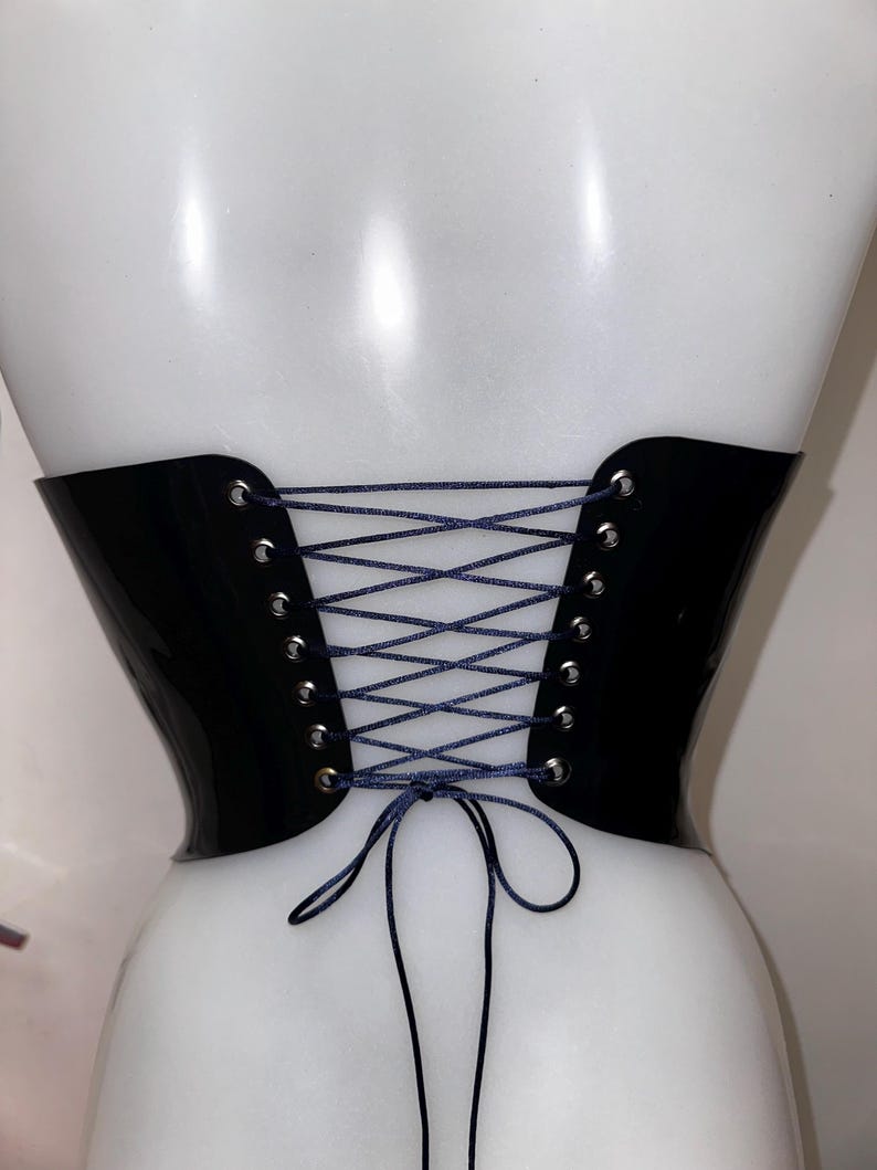 May include: A black corset belt with a glossy finish, featuring silver-toned metal eyelets along the back edges. The garment is secured with a thin, dark blue cord laced in a crisscross pattern and tied in a bow at the bottom centre.