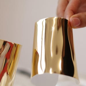 Könnte beinhalten: Zwei goldfarbene Metall-Armreifen. Die Armreifen sind glatt und glänzend und haben eine leicht gebogene Form.