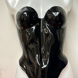 May include: A strapless black corset with a high-gloss, reflective finish. The garment features a sweetheart neckline and a structured, form-fitting silhouette. A small, metallic gold-toned logo is centred on the front of the bodice.