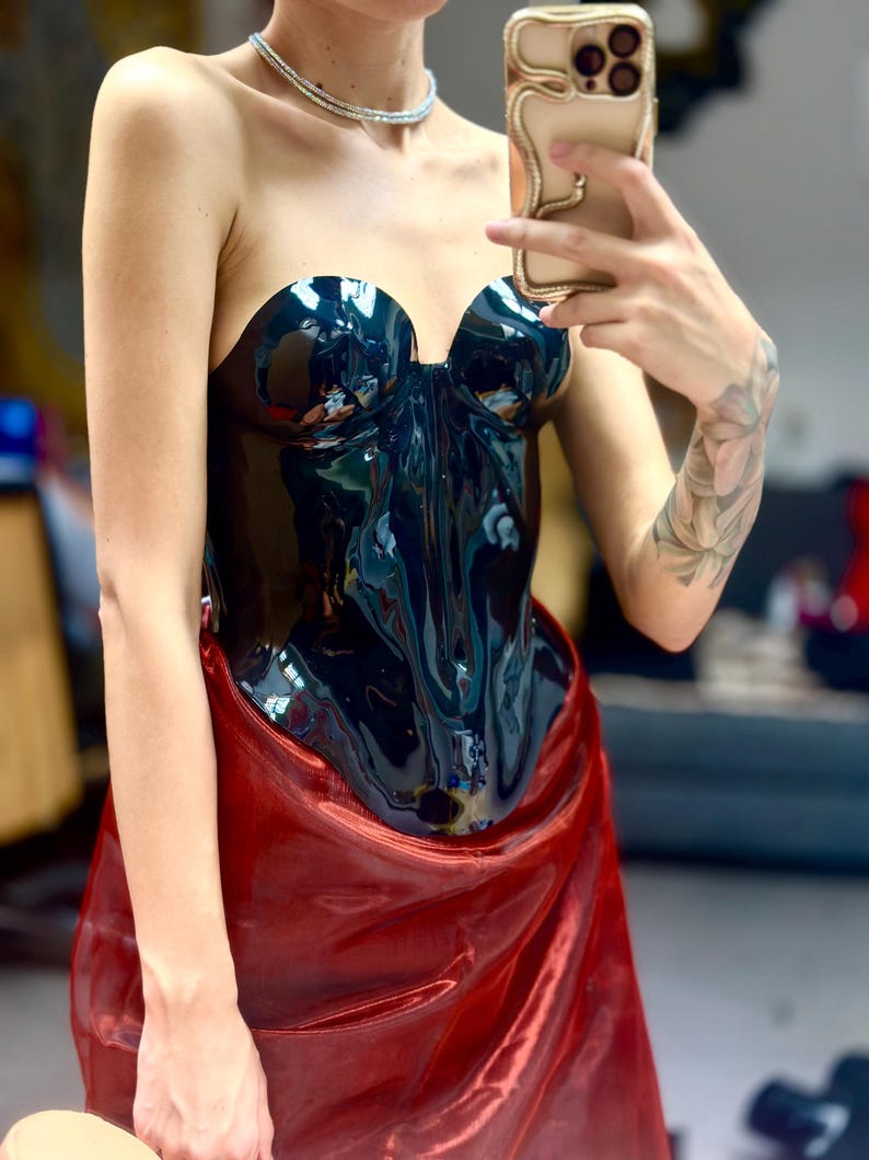 May include: A strapless, high-gloss black corset top with a structured, moulded bodice featuring a sweetheart neckline. The garment is paired with a shimmering, metallic red fabric draped around the waist, creating a distinct contrast in texture.