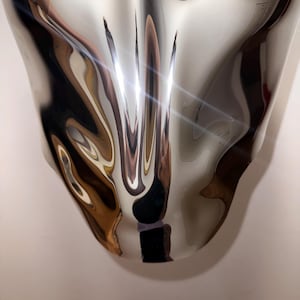 May include: A silver, abstract, metallic sculpture with a smooth, reflective surface. The sculpture is shaped like a teardrop and has a distorted, wavy appearance.