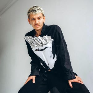 May include: A person wearing a black long-sleeved shirt with a silver reflective design on the front. The shirt is tucked into black pants with a sheer overlay.