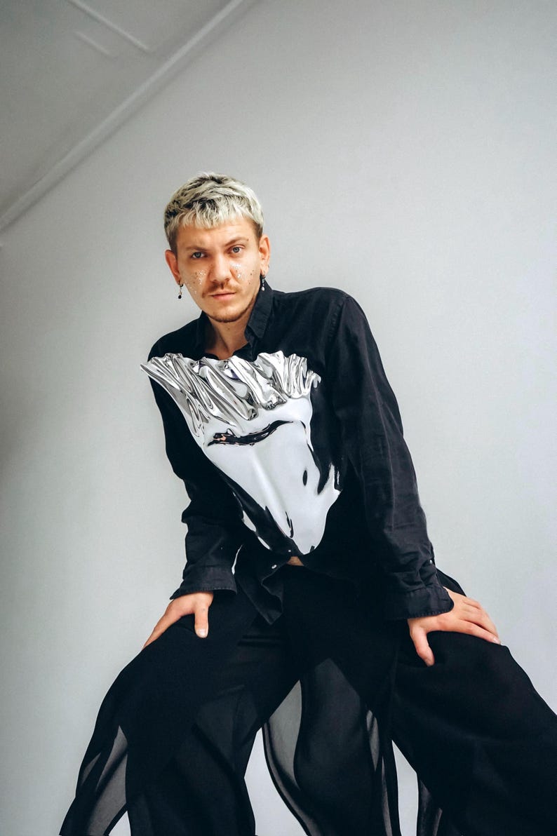 May include: A person wearing a black long-sleeved shirt with a silver reflective design on the front. The shirt is tucked into black pants with a sheer overlay.