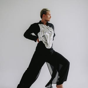 May include: A person wearing a black shirt and pants with a silver metallic corset. The person is standing with one leg forward and is looking to the right.