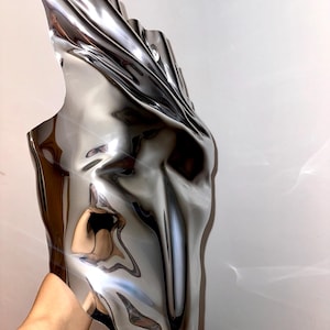 May include: A silver, abstract, metallic sculpture with a smooth, reflective surface. The sculpture is shaped like a face with a long, narrow nose and a wide, open mouth.