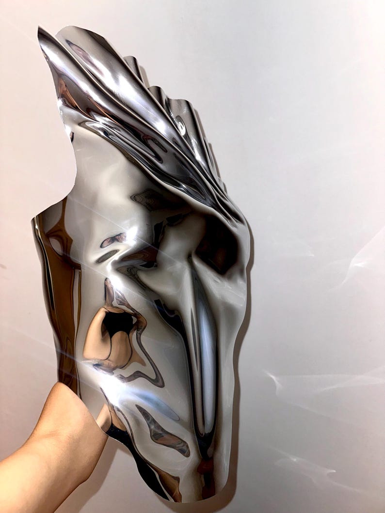 May include: A silver, abstract, metallic sculpture with a smooth, reflective surface. The sculpture is shaped like a face with a long, narrow nose and a wide, open mouth.