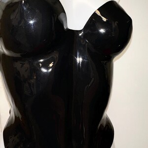 May include: A strapless black bodysuit with a high-gloss, reflective finish. The garment features a sweetheart neckline and a form-fitting silhouette that contours the torso. The material has a smooth, shiny texture that catches light.