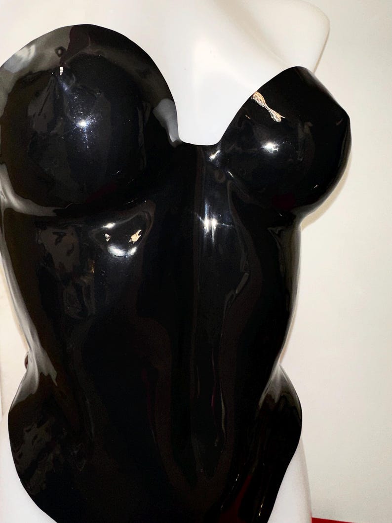 May include: A strapless black bodysuit with a high-gloss, reflective finish. The garment features a sweetheart neckline and a form-fitting silhouette that contours the torso. The material has a smooth, shiny texture that catches light.