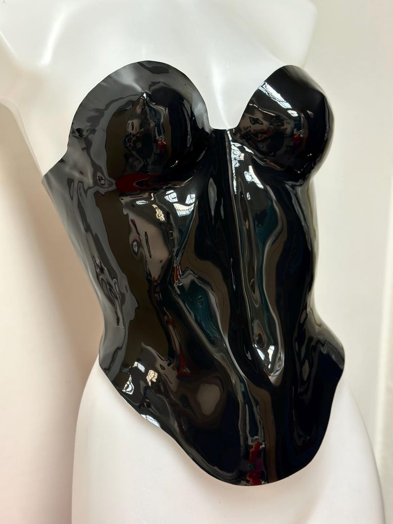 May include: A strapless, glossy black corset top with a moulded bodice and a sweetheart neckline. The garment features a high-shine, reflective finish that contours to the shape of the torso, extending down to a curved hemline at the waist.