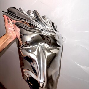 May include: A silver abstract sculpture with a smooth, reflective surface. The sculpture is shaped like a face with a long, pointed nose and a wide mouth.